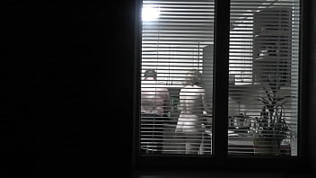 espiando Voyeur. Un vecino voyeur pervertido por la noche en la calle mira por la ventana mientras un vecino desnudo y sexy en la cocina prepara la cena para su marido. Desnudo en público. Desnuda en casa. Familia. Exterior